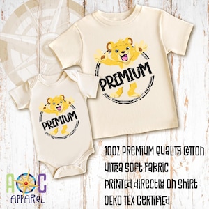 May include: A cream-colored short-sleeve t-shirt and a matching short-sleeve bodysuit, both featuring a graphic of a yellow lion cub inside a black circular frame with the text PREMIUM. Text on the image reads 100% PREMIUM QUALITY COTTON.