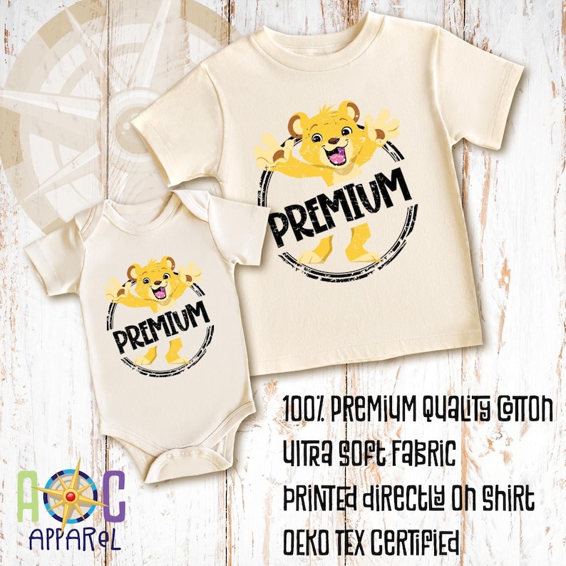 May include: A cream-colored short-sleeve t-shirt and a matching short-sleeve bodysuit, both featuring a graphic of a yellow lion cub inside a black circular frame with the text PREMIUM. Text on the image reads 100% PREMIUM QUALITY COTTON.