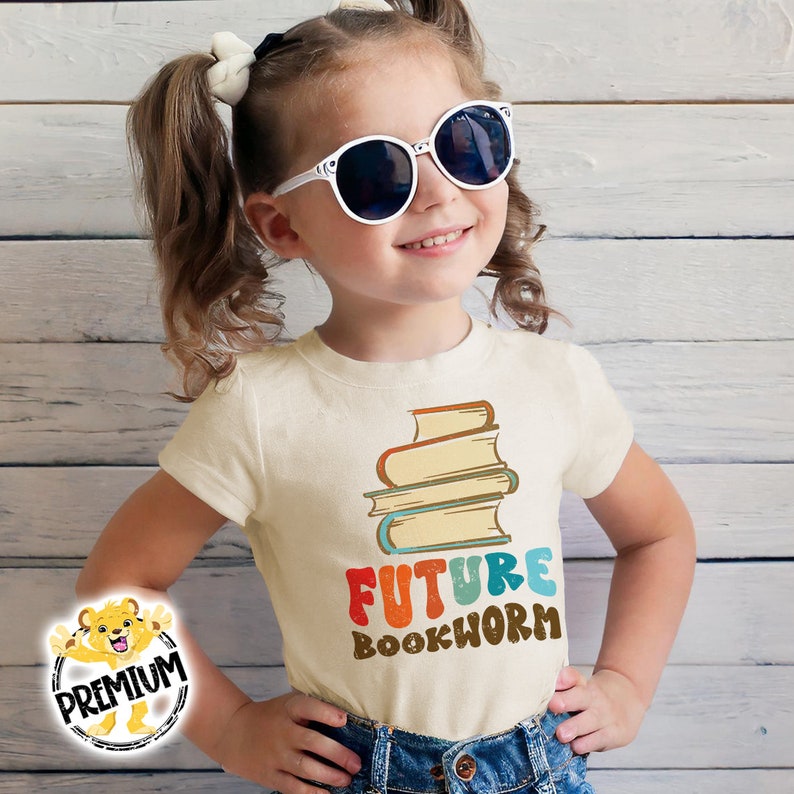 Pu&ograve; includere: Una maglietta bianca con una pila di libri colorati e il testo "FUTURE BOOKWORM" in un font retr&ograve;.