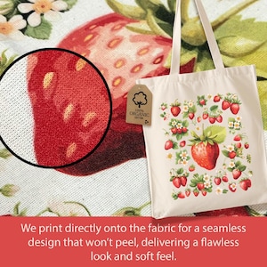 May include: A white canvas tote bag with a design of red strawberries and white flowers. The bag has two handles and a label that says "100% Organic Cotton".