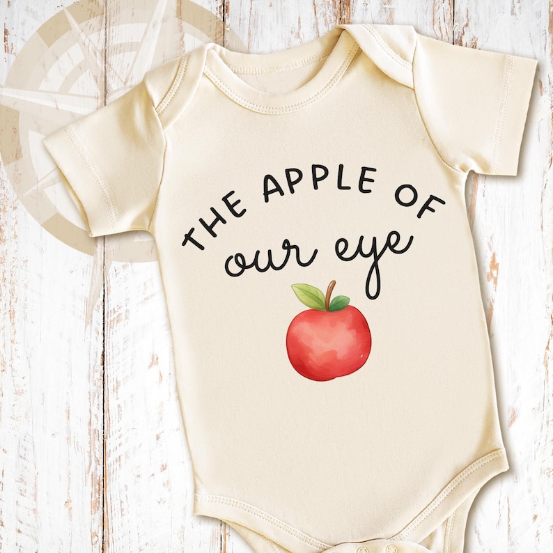 May include: A cream-colored short-sleeve bodysuit featuring the black text "THE APPLE OF our eye" printed above a watercolor-style illustration of a red apple with a green leaf and a brown stem centered on the front of the garment.