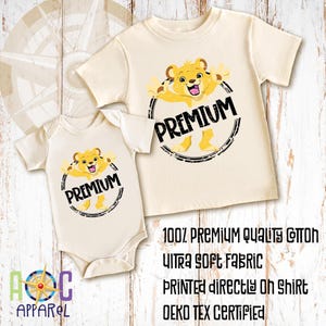 May include: A cream-colored short-sleeve t-shirt and a matching short-sleeve bodysuit, both featuring a graphic of a yellow lion cub inside a black circular frame with the text PREMIUM. Text on the image reads 100% PREMIUM QUALITY COTTON.