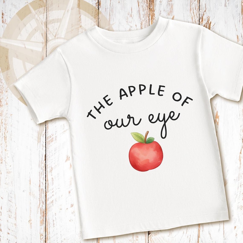 May include: A white short-sleeved t-shirt featuring the black printed text THE APPLE OF our eye centered on the chest. Below the text is a watercolor-style illustration of a single red apple with a green leaf and a brown stem.