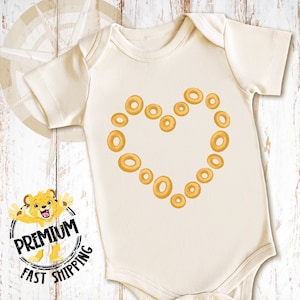 May include: A white baby onesie with a heart shape made of yellow cereal rings.