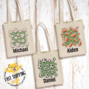 May include: Three beige canvas tote bags with sports ball designs. One bag features baseballs, another footballs, and the third soccer balls. Each bag has a name printed below the design: Michael, Aiden, and Daniel.