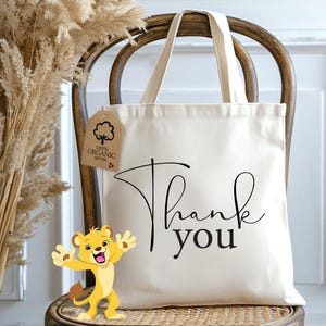 May include: A white canvas tote bag with black text that says "Thank you". The bag has a brown tag attached to it that says "100% Organic Cotton". A cartoon lion is in the foreground.