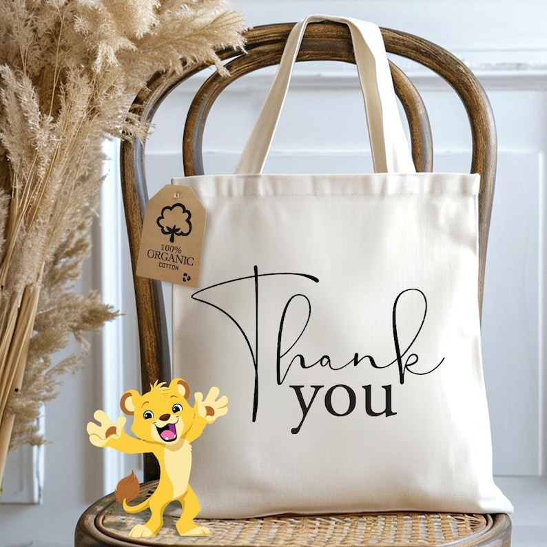 May include: A white canvas tote bag with black text that says "Thank you". The bag has a brown tag attached to it that says "100% Organic Cotton". A cartoon lion is in the foreground.