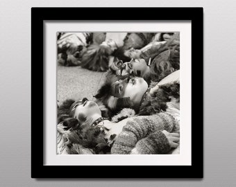 May include: Black and white photograph of a group of people lying down with their faces turned upwards. They are wearing costumes with feathers and fur. The image is framed in a black frame.