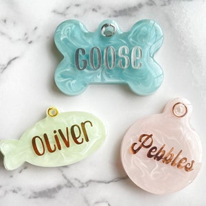 May include: Three pet ID tags in the image. The top tag is bone-shaped, light blue with the name "GOOSE" in silver. The middle tag is fish-shaped, light green with "Oliver" in gold. The bottom tag is round, pink with "Pebbles" in gold.