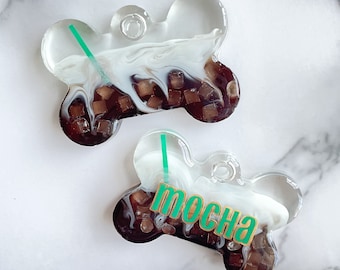 Peut inclure: Deux étiquettes pour animaux de compagnie en forme d'os avec un motif moka. Les étiquettes sont transparentes avec un motif tourbillonnant blanc et marron, des cubes bruns et une paille verte. Une étiquette porte le mot "mocha" en vert.