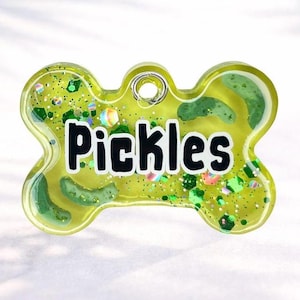 May include: A bone-shaped pet tag with the name "Pickles" in bold black letters. The tag is filled with a yellow-green liquid, pickle slices, and colorful glitter. A small metal ring is at the top for attachment.