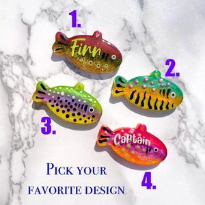 Pu&ograve; includere: Quattro ciondoli in resina a forma di pesce con accenti glitterati. Ogni ciondolo ha un design unico, inclusi nomi come "Finn" e "Captain". &Egrave; presente anche il testo "Pick your favorite design". Questi ciondoli sono probabilmente per gioielli o artigianato.