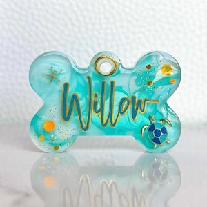 May include: A turquoise and white dog tag in a bone shape, with the name "Willow" in gold script. The tag features a small turtle, starfish, and gold glitter accents. A gold-colored ring is at the top for attachment.