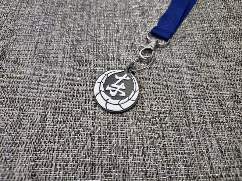 May include: A silver key chain with a black and white circular charm featuring a Japanese character. The charm is attached to a blue lanyard.
