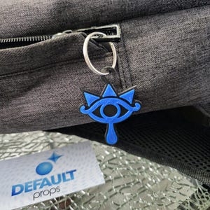 May include: A blue and black eye symbol keychain with a silver ring. The keychain is attached to a gray backpack.