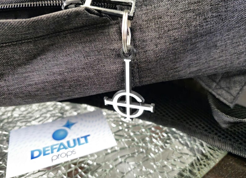 May include: A silver keychain featuring a black and white inverted pentagram symbol. The keychain is attached to a black zipper pull on a grey bag.