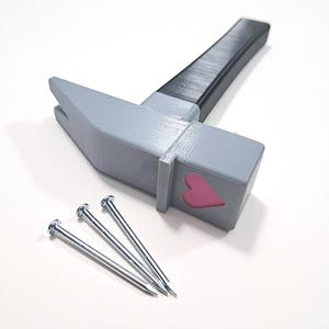 May include: A gray hammer with a black handle and a pink heart design on the head. Three silver nails are lying next to the hammer.