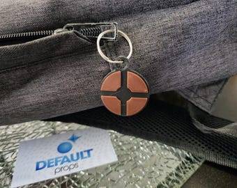 May include: A brown and black circular keychain with a symbol resembling a target. The keychain is attached to a silver key ring and is hanging from a zipper on a gray backpack.