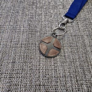 Puede incluir: Un llavero de metal de color plateado y cobre con un dise&ntilde;o circular que presenta una estrella de cuatro puntas. El llavero est&aacute; unido a un cord&oacute;n azul.