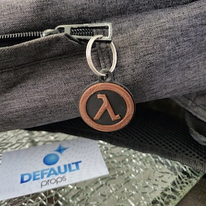 May include: A black and brown 3D printed keychain with the Half-Life logo. The keychain is attached to a silver key ring.
