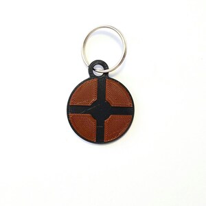 Puede incluir: Un llavero circular negro y marr&oacute;n con un s&iacute;mbolo que se asemeja a una cruz con cuatro extremos redondeados. El llavero tiene un anillo de metal plateado.