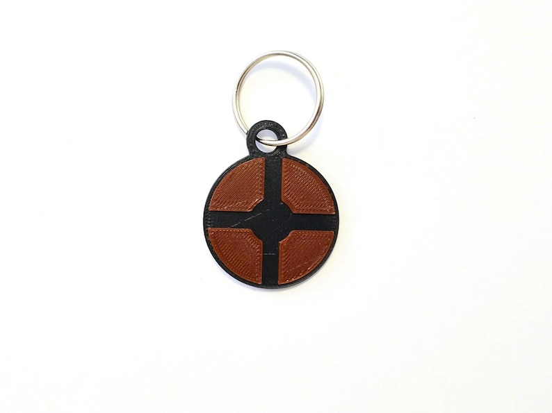 Puede incluir: Un llavero circular negro y marr&oacute;n con un s&iacute;mbolo que se asemeja a una cruz con cuatro extremos redondeados. El llavero tiene un anillo de metal plateado.