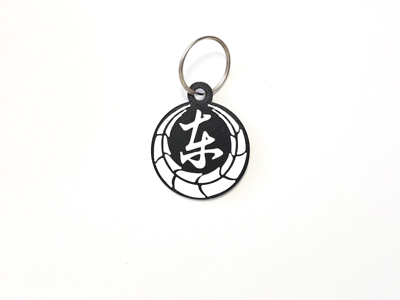 May include: Black and white keychain with a circle design featuring a Japanese character in the center. The character is "老" which means "old" or "elder".