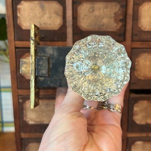 以下が含まれることがあります： ファセットデザインの透明なガラス製のドアノブ。ノブは手に持っており、複数の引き出しが付いた木製のキャビネットの前にある。