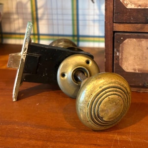 Op de afbeelding: Een vintage messing deurknop met een rond, gegroefd ontwerp. De knop is bevestigd aan een zwart metalen deurgreep met een vergrendeling.