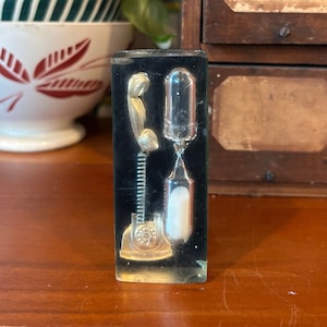 May include: A clear glass hourglass with a black frame and a gold-coloured phone receiver. The hourglass is filled with white sand and is sitting on a wooden surface.