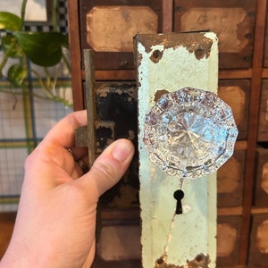 May include: A vintage door knob with a clear glass crystal knob and a keyhole. The knob is attached to a metal plate with a light green paint finish. The plate has some rust and wear.