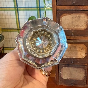 以下が含まれることがあります： 装飾的なデザインの、透明でファセット加工されたガラス製のドアノブ。ノブは、小さな引き出しが付いたヴィンテージの木製キャビネットの前に保持されており、家の装飾と修復に焦点を当てていることを示唆しています。