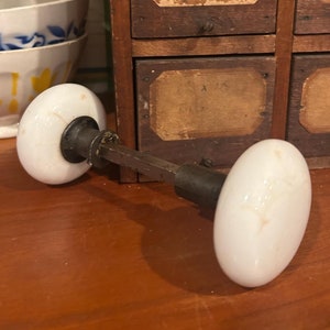 Peut inclure: Une paire de boutons de porte en porcelaine blanche avec une tige métallique les reliant. Les boutons sont de forme ovale et ont un design simple. La tige est brune et a une finition en laiton.