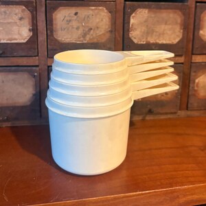 Può includere: Un set di cinque misurini in plastica color bianco sporco impilati verticalmente. Ogni misurino presenta una coppa rotonda e un manico piatto sporgente. I misurini sono inseriti l'uno nell'altro, creando un aspetto a gradini.