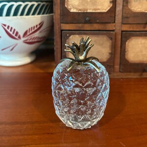 May include: A clear glass pineapple shaped decorative object with a gold metal top. The pineapple is sitting on a wooden surface.