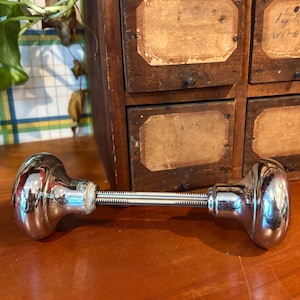 Puede incluir: Un pomo de puerta cromado brillante con una varilla roscada larga. El pomo tiene una forma redondeada y bulbosa. El fondo incluye un mueble de madera con pequeños cajones y una planta.