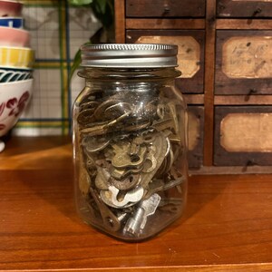 May include: A glass jar filled with a collection of vintage keys. The jar has a silver lid and is sitting on a wooden surface.