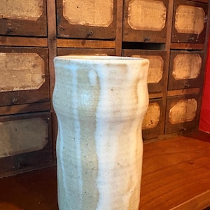 May include: A cylindrical ceramic vase with a textured, off-white and beige glaze. The surface features vertical ridges and a subtle drip pattern where the lighter glaze overlaps the darker base tone, creating a varied, earthy appearance.