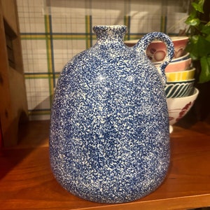 Puede incluir: Una jarra de cerámica azul y blanca con un esmalte moteado. La jarra tiene un cuerpo redondo y un asa.