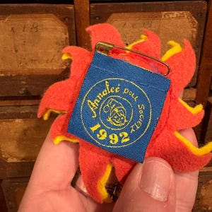 Puede incluir: Un broche en forma de flor roja y amarilla con una etiqueta azul adjunta. La etiqueta tiene texto dorado y el a&ntilde;o "1992" impreso. Un imperdible dorado asegura la etiqueta a la flor.