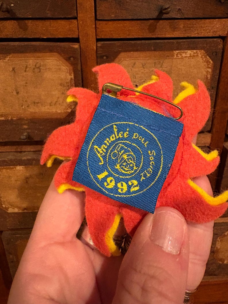 Puede incluir: Un broche en forma de flor roja y amarilla con una etiqueta azul adjunta. La etiqueta tiene texto dorado y el a&ntilde;o "1992" impreso. Un imperdible dorado asegura la etiqueta a la flor.