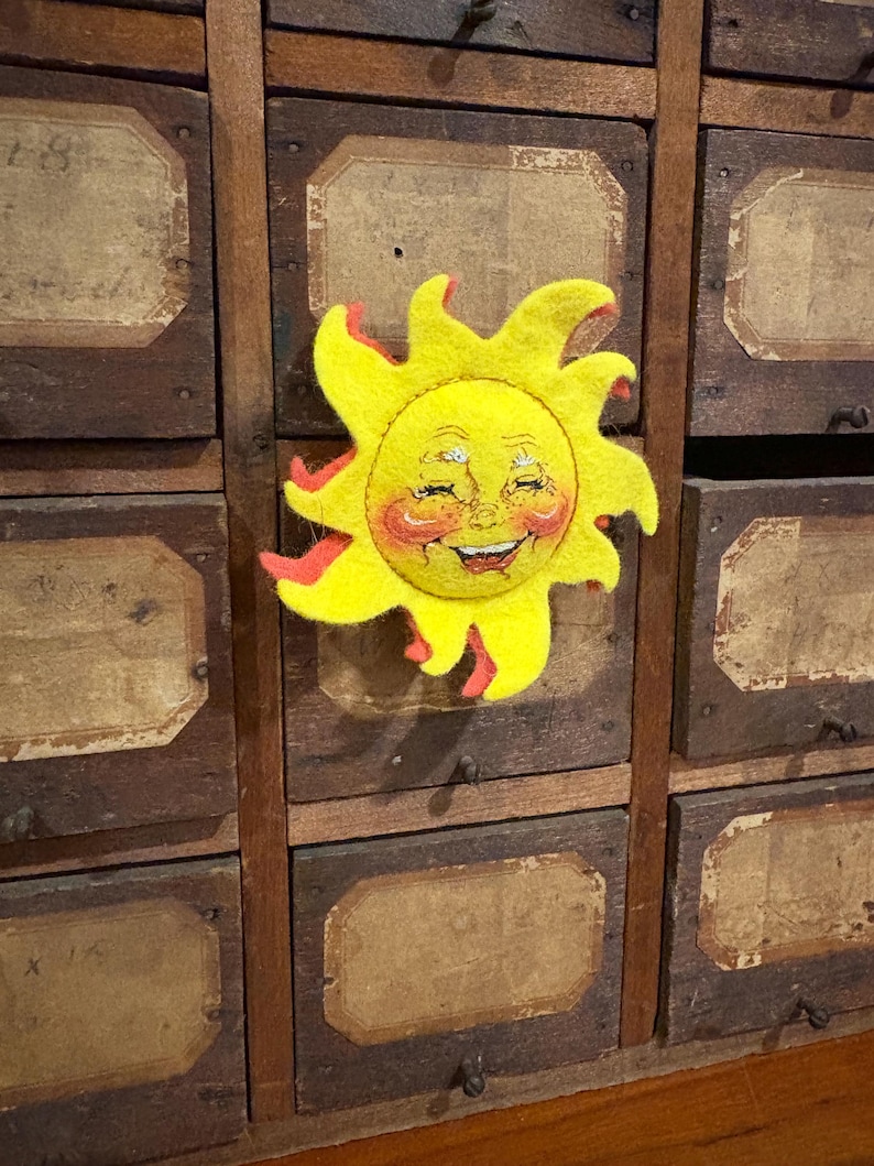 Puede incluir: Un alegre sol de fieltro hecho a mano con una cara sonriente, mejillas rosadas y detalles rojos. El sol amarillo est&aacute; fijado a un mueble de madera vintage con m&uacute;ltiples cajones, cada uno con una etiqueta. El dise&ntilde;o del sol evoca una est&eacute;tica artesanal caprichosa.
