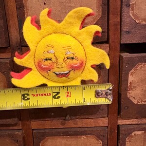 Puede incluir: Un sol de fieltro con una cara sonriente y detalles rojos. El sol es amarillo con una cara sonriente, mejillas rosadas y llamas rojas. El sol mide aproximadamente 7,6 cm de di&aacute;metro.