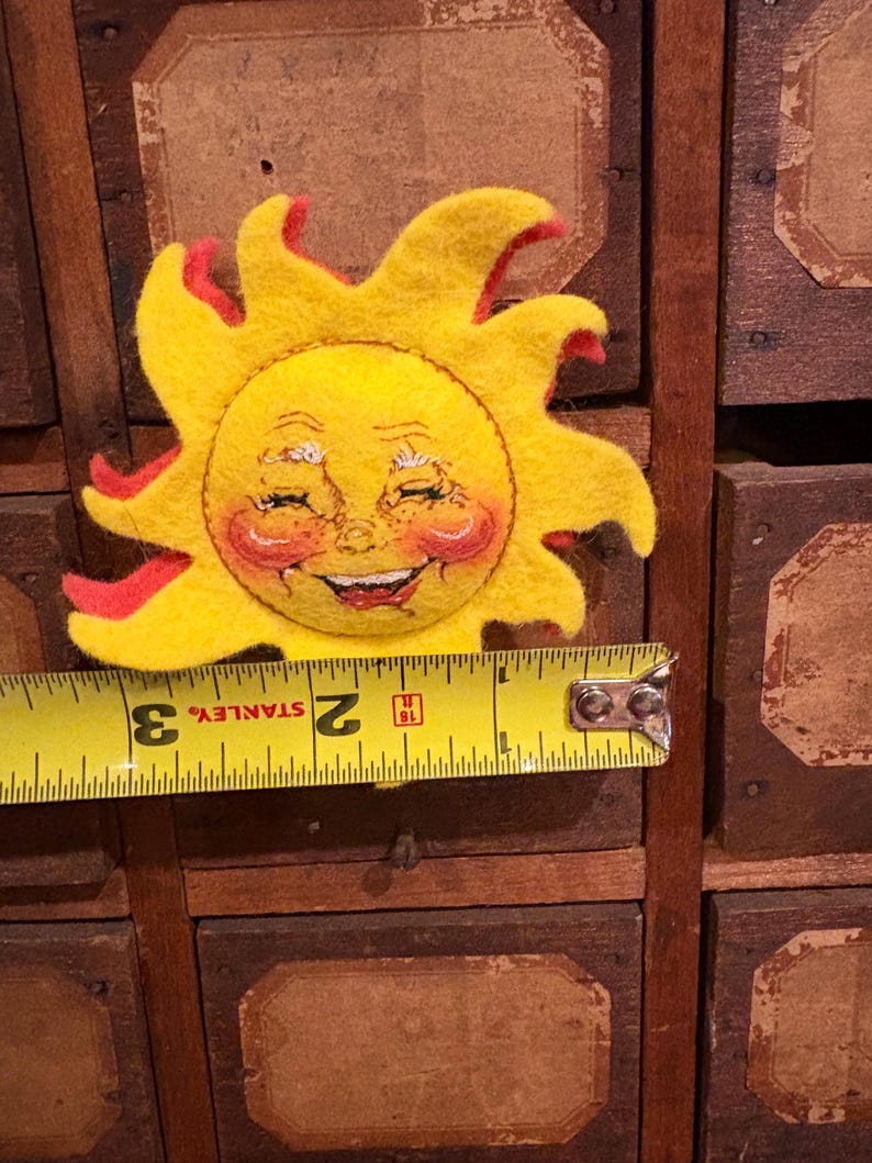 Puede incluir: Un sol de fieltro con una cara sonriente y detalles rojos. El sol es amarillo con una cara sonriente, mejillas rosadas y llamas rojas. El sol mide aproximadamente 7,6 cm de di&aacute;metro.