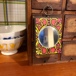 May include: A small, rectangular mirror with a decorative frame. The frame features a red background with yellow and black floral designs. A silver ring is attached to the top for hanging. The mirror is set against a wooden cabinet.