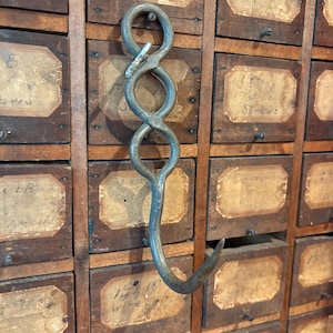 May include: A vintage metal hook with a unique, intertwined design, resembling a figure eight. The hook is mounted on a wooden cabinet with numerous small drawers, each labeled with handwritten text on aged paper.
