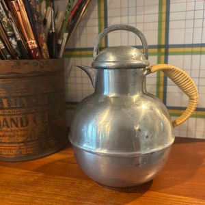 May include: A silver pitcher with a woven handle. The pitcher has a round body and a lid with a knob on top. The pitcher is sitting on a wooden surface.