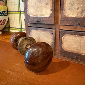 May include: Three antique wooden drawer pulls with a glossy, dark brown finish are arranged on a wooden surface. A vintage wooden cabinet with label holders is in the background. The pulls have a rounded, oval shape.