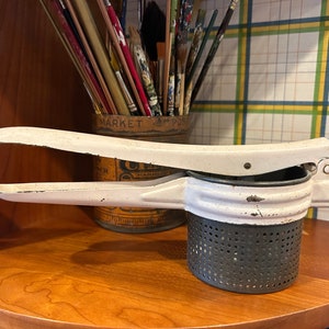 Puede incluir: Prensador de patatas de metal blanco con un colador de metal negro. El prensador tiene un mango que está doblado en un ángulo de 90 grados.