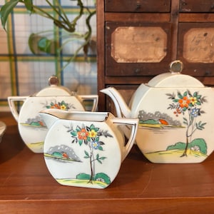 May include: A vintage tea set with a white background and floral designs. The set includes a teapot, creamer, and sugar bowl. The teapot has a round body and a spout on the side. The creamer and sugar bowl have a round body and a handle on the side.